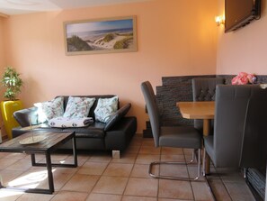 A living room with a sofa, a dining table, and a chair. There is a painting on the wall and a potted plant.