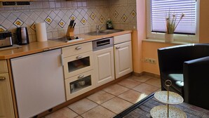 A kitchen with white cabinets, a tiled backsplash, a window with blinds, and a small table with decorative items. - Kos - sparrow lust - sparrow lust (Koserow)