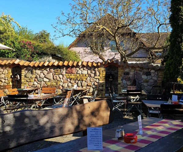 An outdoor seating area with stone walls and wooden furniture. Tables are set up with chairs, and a menu sign is visible.