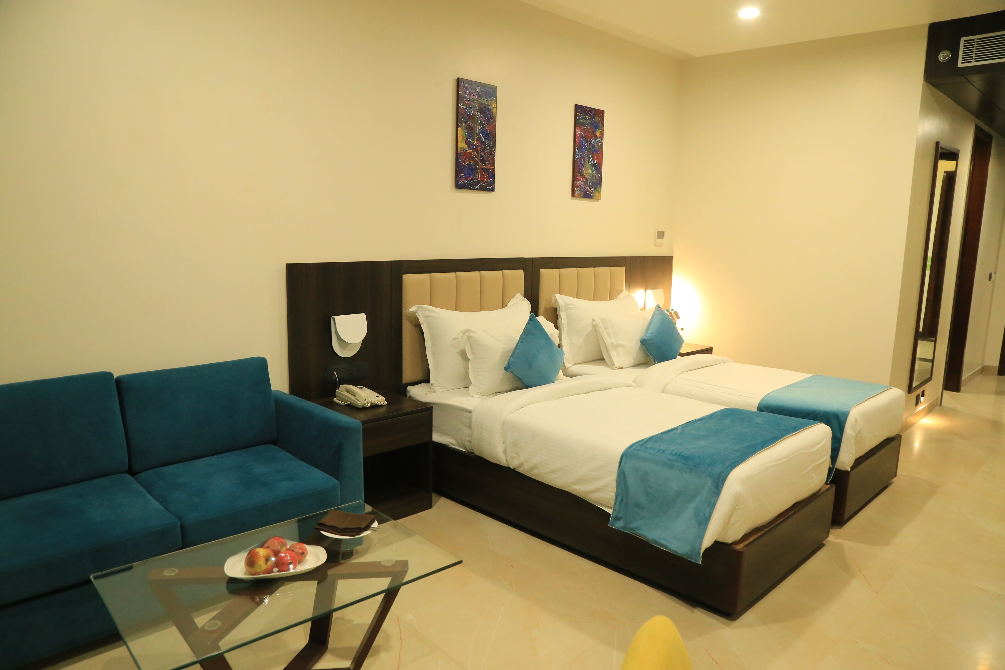 Habitación de hotel con sofá, dos camas, una mesa de centro de vidrio con fruta y obras de arte en las paredes.