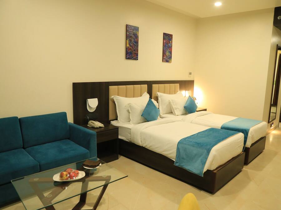 Habitación de hotel con sofá, dos camas, una mesa de centro de vidrio con fruta y obras de arte en las paredes.