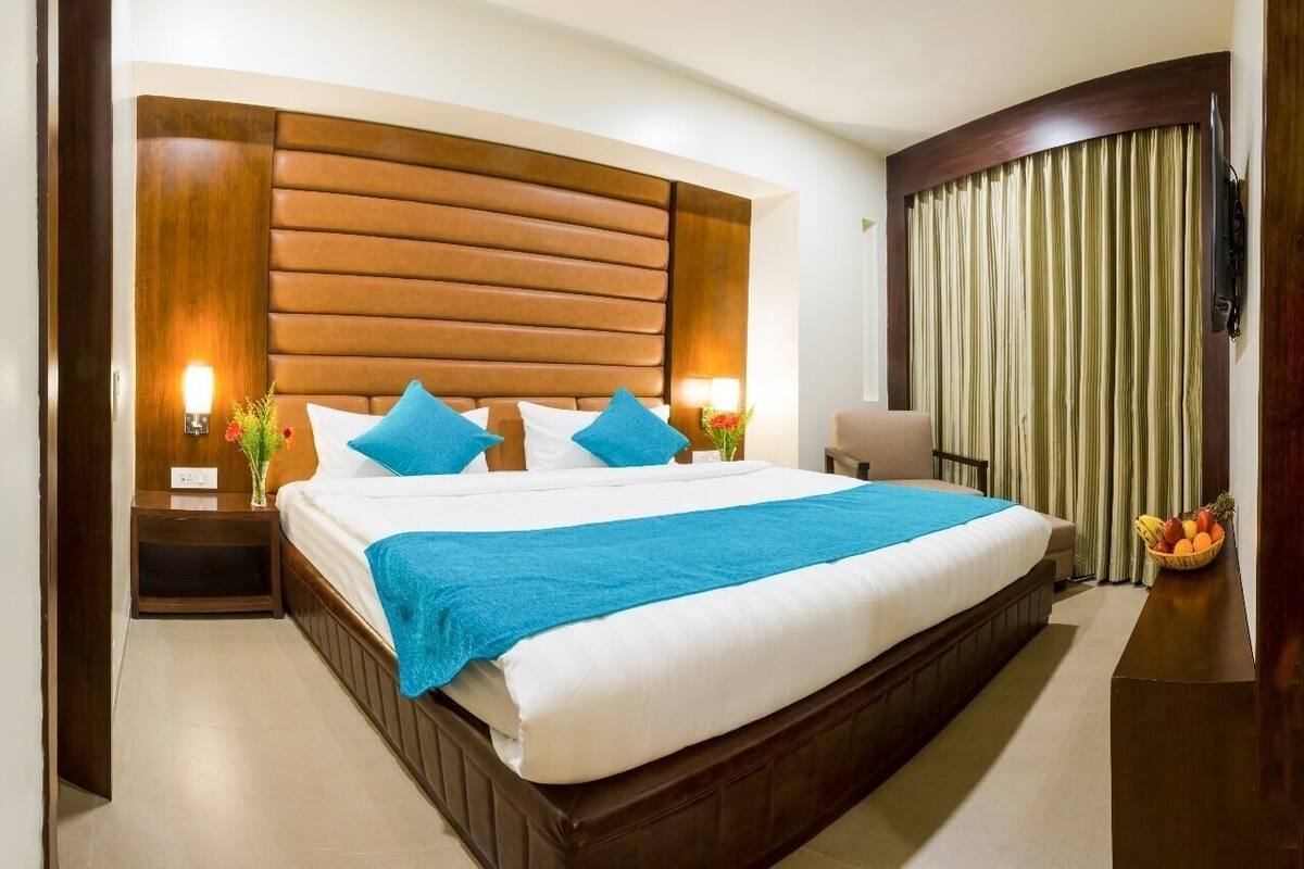 Quarto de hotel com uma cama grande, cabeceira de madeira, duas luminárias de mesa, mesa de cabeceira, uma cadeira, televisão e cortina.