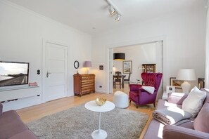 A modern living room with a sofa, armchair, and coffee table. There is a television on a stand, a dresser, and a dining table with chairs. - The locksmith's shop, Germany (Sylt)