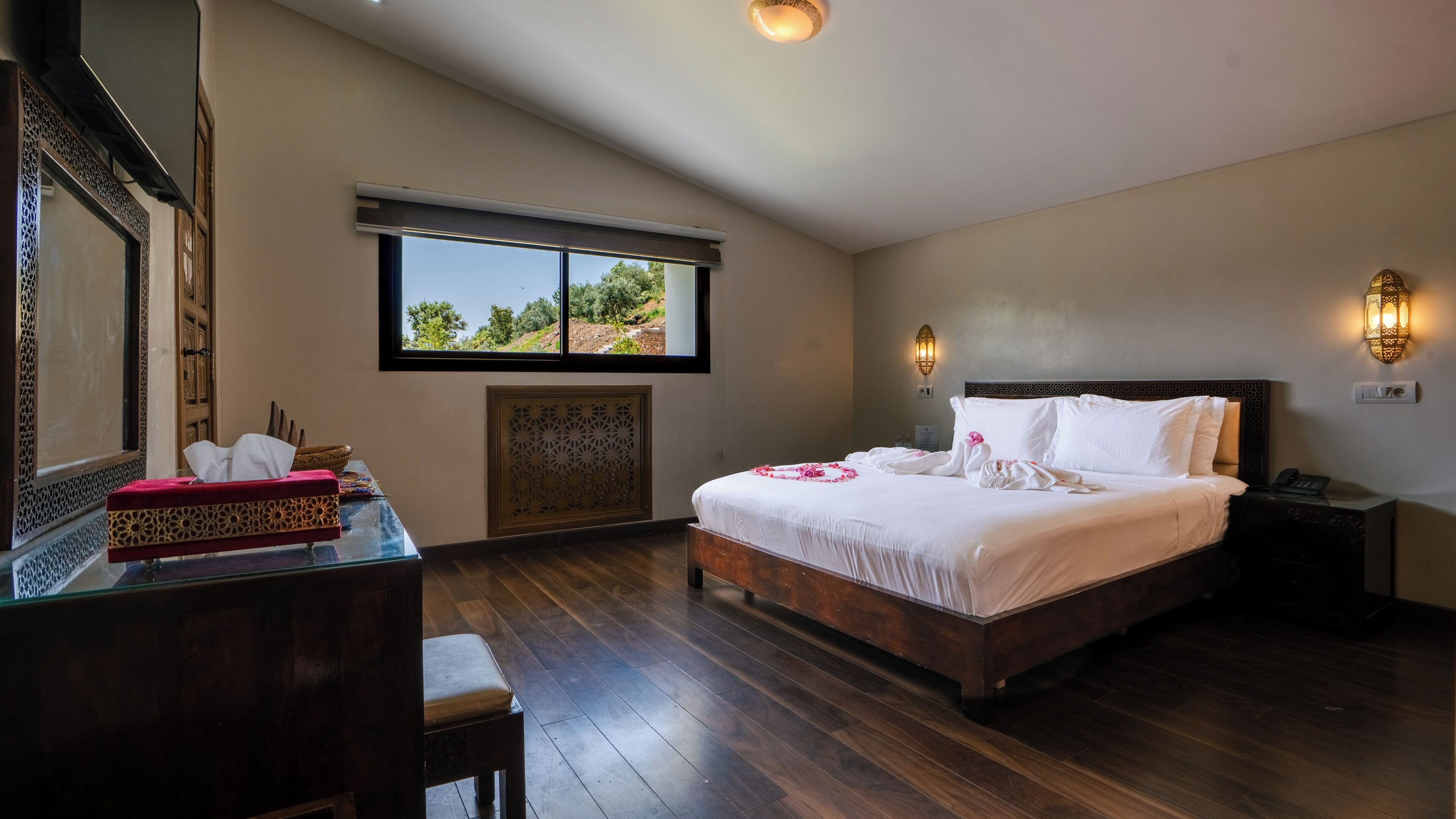 A hotel room with a large bed, a desk with a red cloth, a window with a view of greenery, and a wall-mounted light fixture.