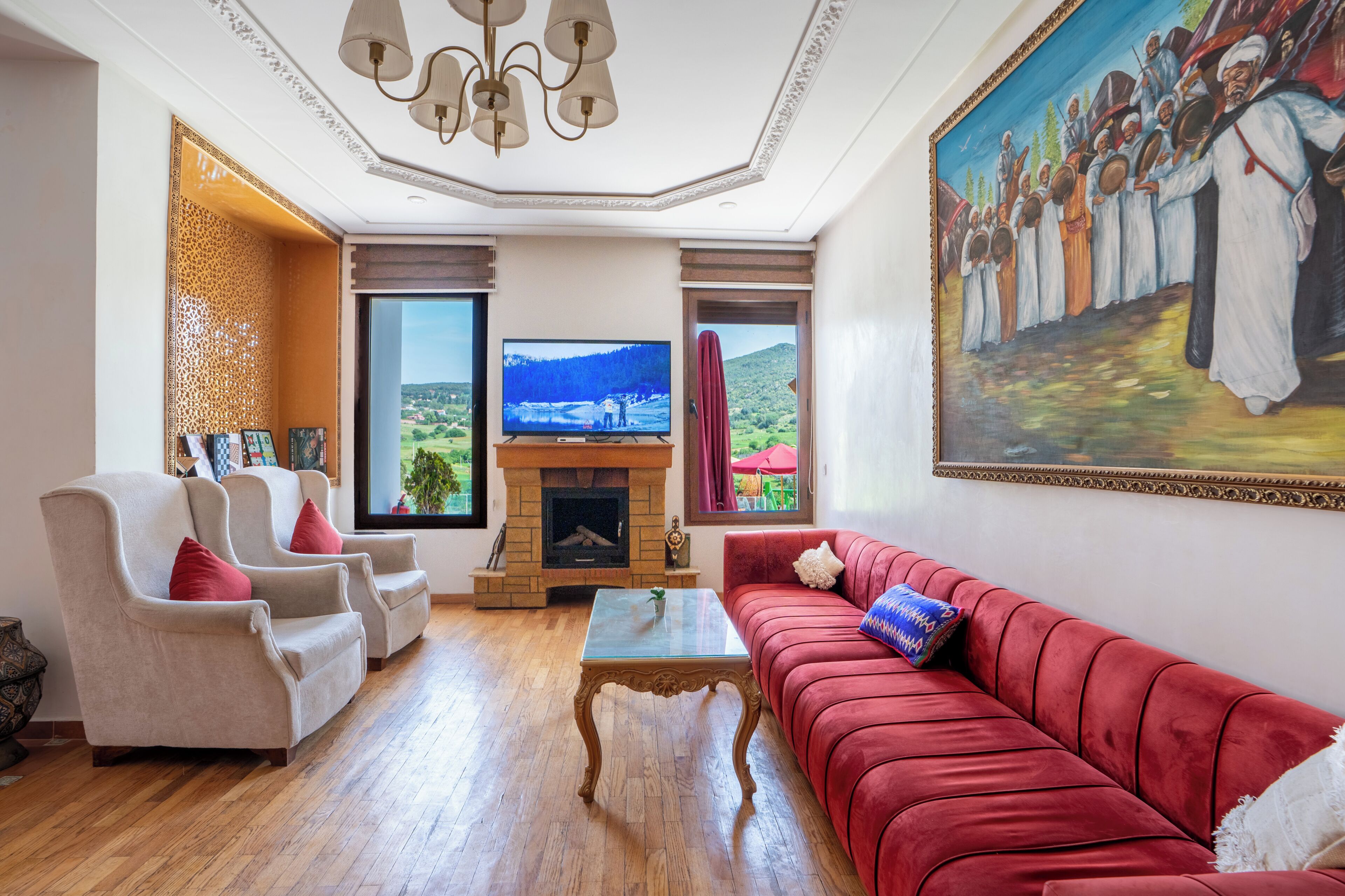 Een woonkamer met een open haard, een rode bank, een beige fauteuil en een televisie.