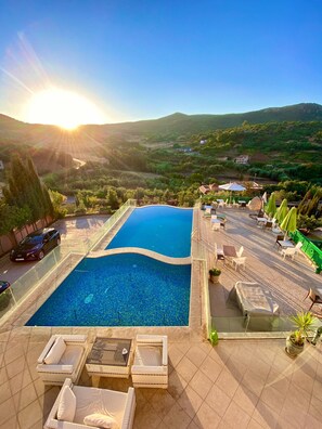 Una piscina con sillones y mesas, rodeada por un patio con vistas a un paisaje montañoso.
