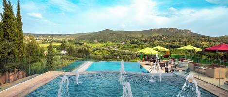 Una piscina infinita con chorros de agua, rodeada de asientos exteriores y sombrillas, con vistas a un paisaje montañoso.