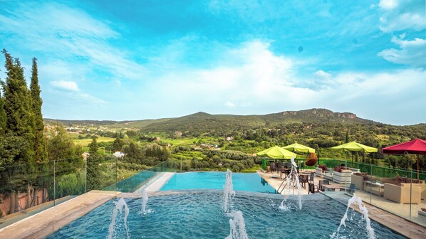 An infinity pool with water jets, surrounded by outdoor seating and umbrellas, overlooking a mountainous landscape.