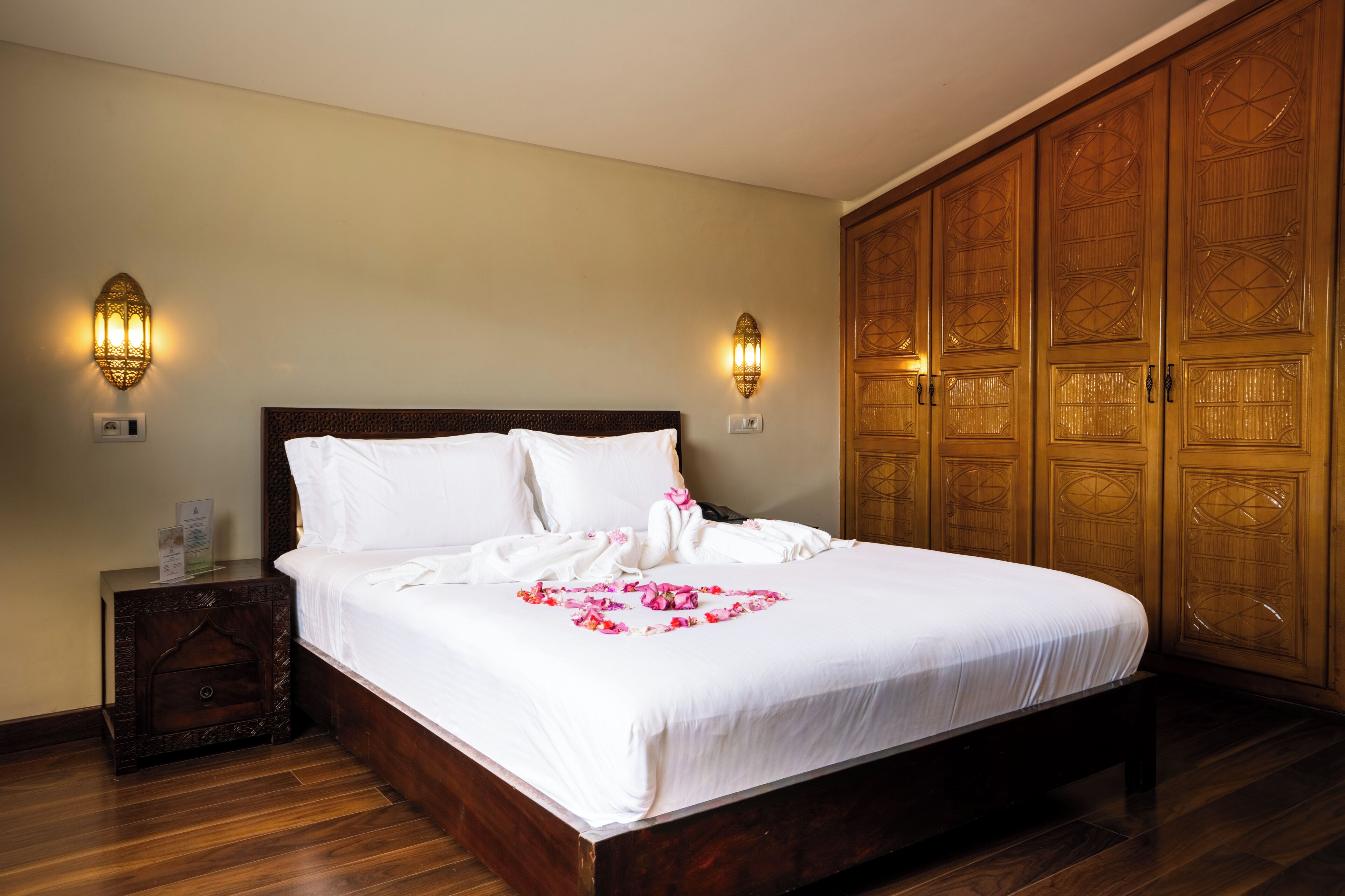 A hotel room with a large bed, wooden wardrobe, two wall sconces, and a nightstand.