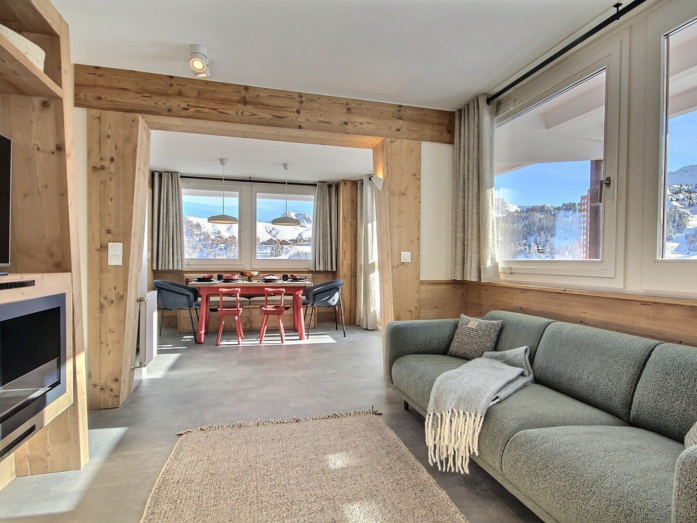 Une pièce chaleureuse avec un plafond en bois, un canapé, une télévision, une table à manger avec des chaises et de grandes fenêtres offrant une vue sur les montagnes.