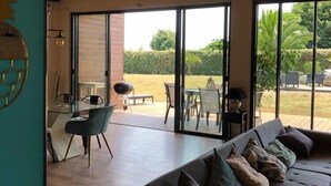 A modern living room with a large sofa, a dining area with a table and chairs, and a view of a garden through large windows. - VILLA 260€ 12 People Ideal for large groups (hikers, families, etc.) (Plouenan ( kerlaudy))