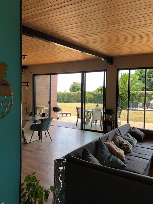 Una sala de estar moderna con un sofá grande, un comedor con mesa y sillas, y vistas a un jardín a través de amplios ventanales.