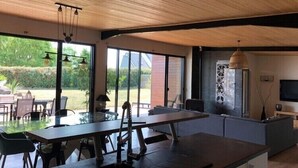 A modern kitchen with a large island, a dining area with a glass table, and a view of the garden through large windows. - VILLA 260€ 12 People Ideal for large groups (hikers, families, etc.) (Plouenan ( kerlaudy))
