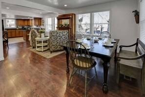 On aperçoit un coin repas avec une table en bois dressée pour quatre personnes, entourée de chaises à motifs. Au fond, on distingue une cuisine avec des placards en bois et une fenêtre.