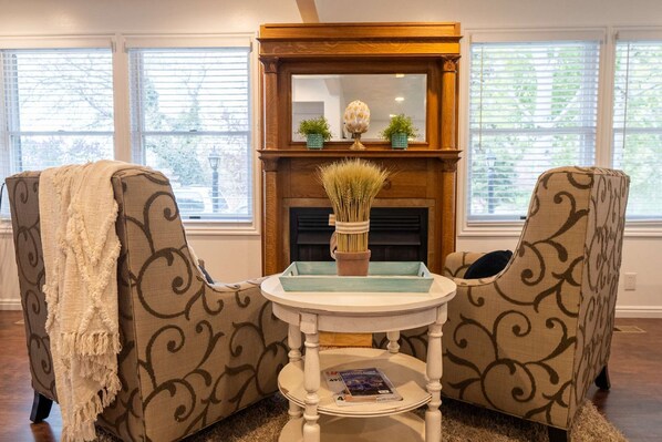 Un salon chaleureux avec deux fauteuils à motifs, une petite table ronde et une cheminée agrémentée d’objets décoratifs.