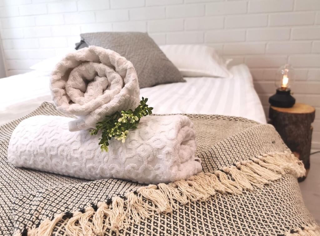 Un lit bien fait, avec des serviettes pliées en rouleau et une petite plante, un plaid à motifs et une table de chevet sur laquelle est posée une bougie.