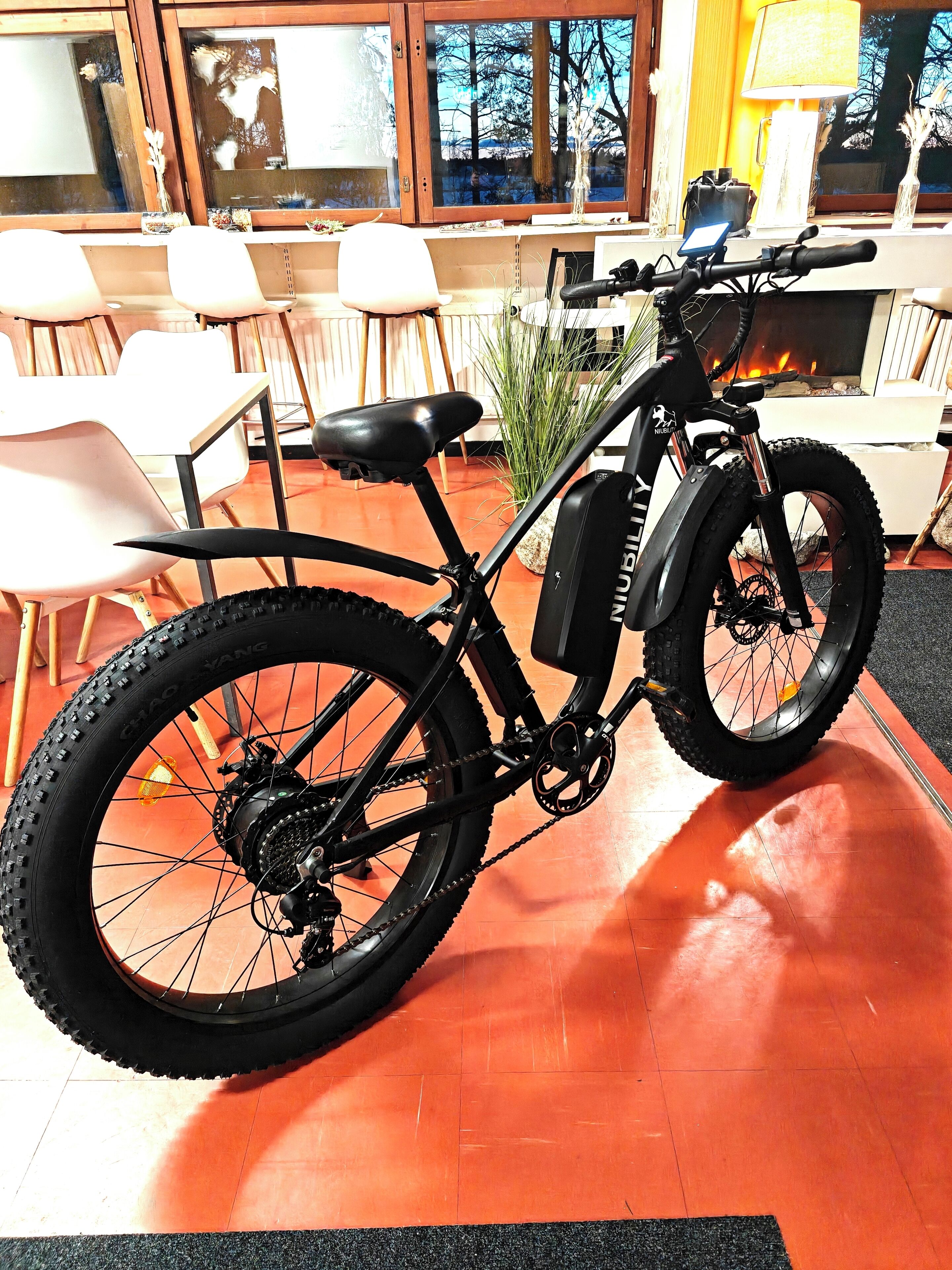 A black electric bike with a seat and front suspension, a chain drive, and thick tires, placed indoors on a red floor with a window in the background.