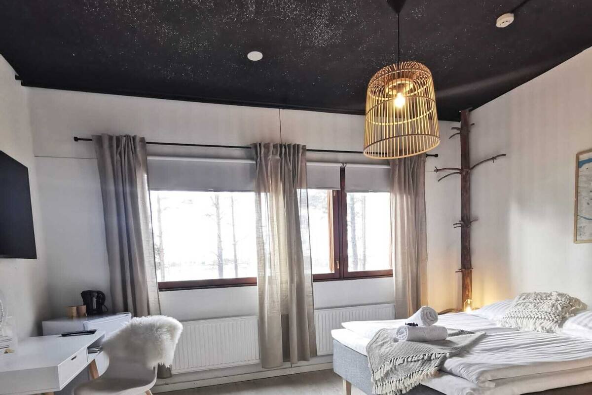 A modern bedroom with a bed, a desk, and a chair. There are windows with curtains, a hanging light fixture, and a radiator.
