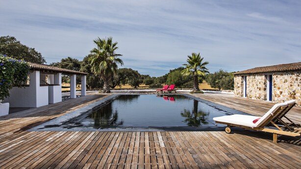 Une terrasse en bois avec une piscine, deux chaises longues rouges et, au fond, des bâtiments en pierre.