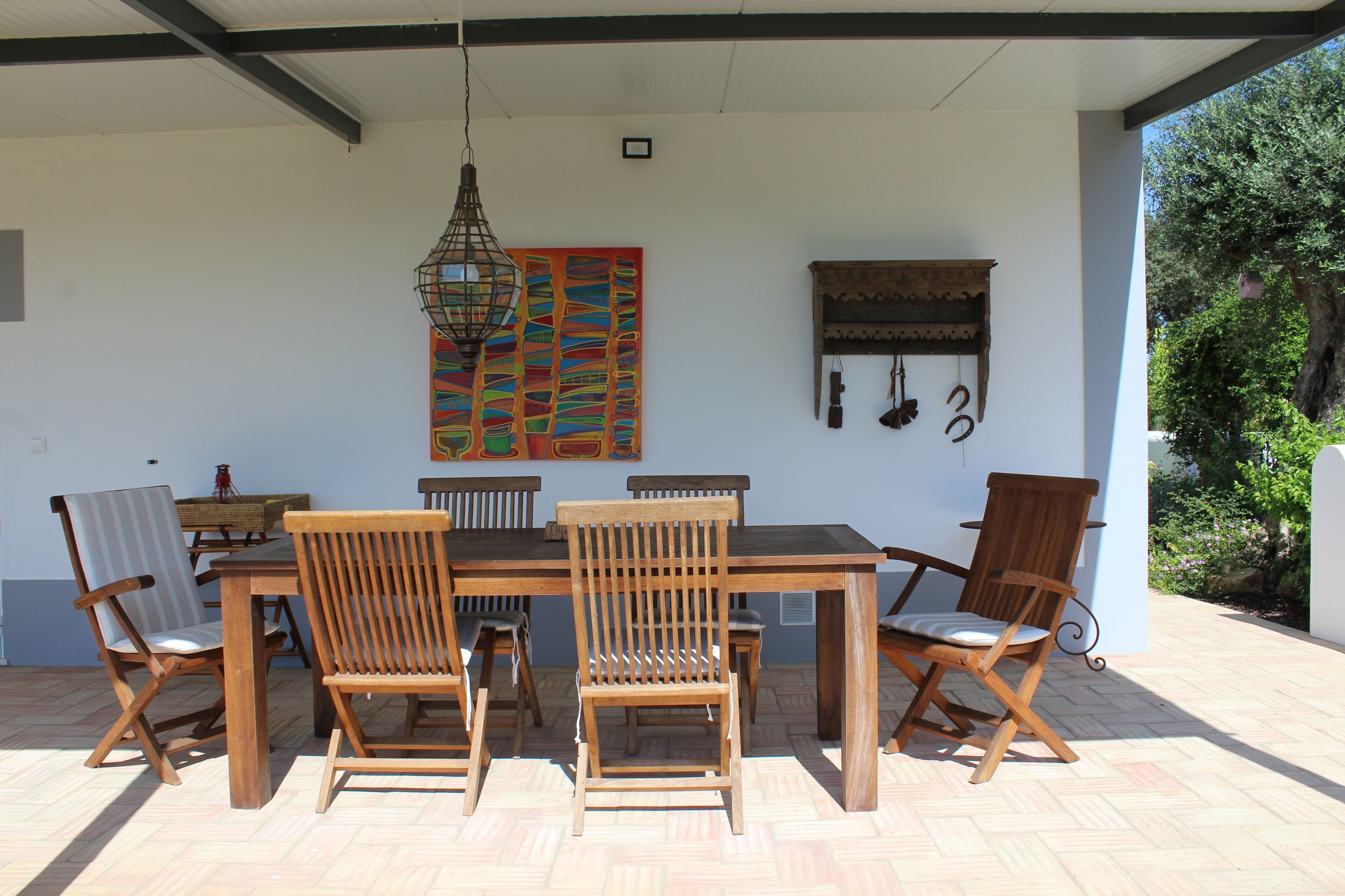 Une terrasse couverte avec une table et des chaises en bois, un luminaire suspendu et une peinture abstraite aux couleurs vives accrochée au mur.