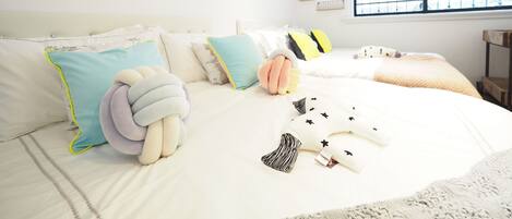 Quarto bem arrumado com uma cama grande, várias almofadas, um brinquedo macio e quadros nas paredes.