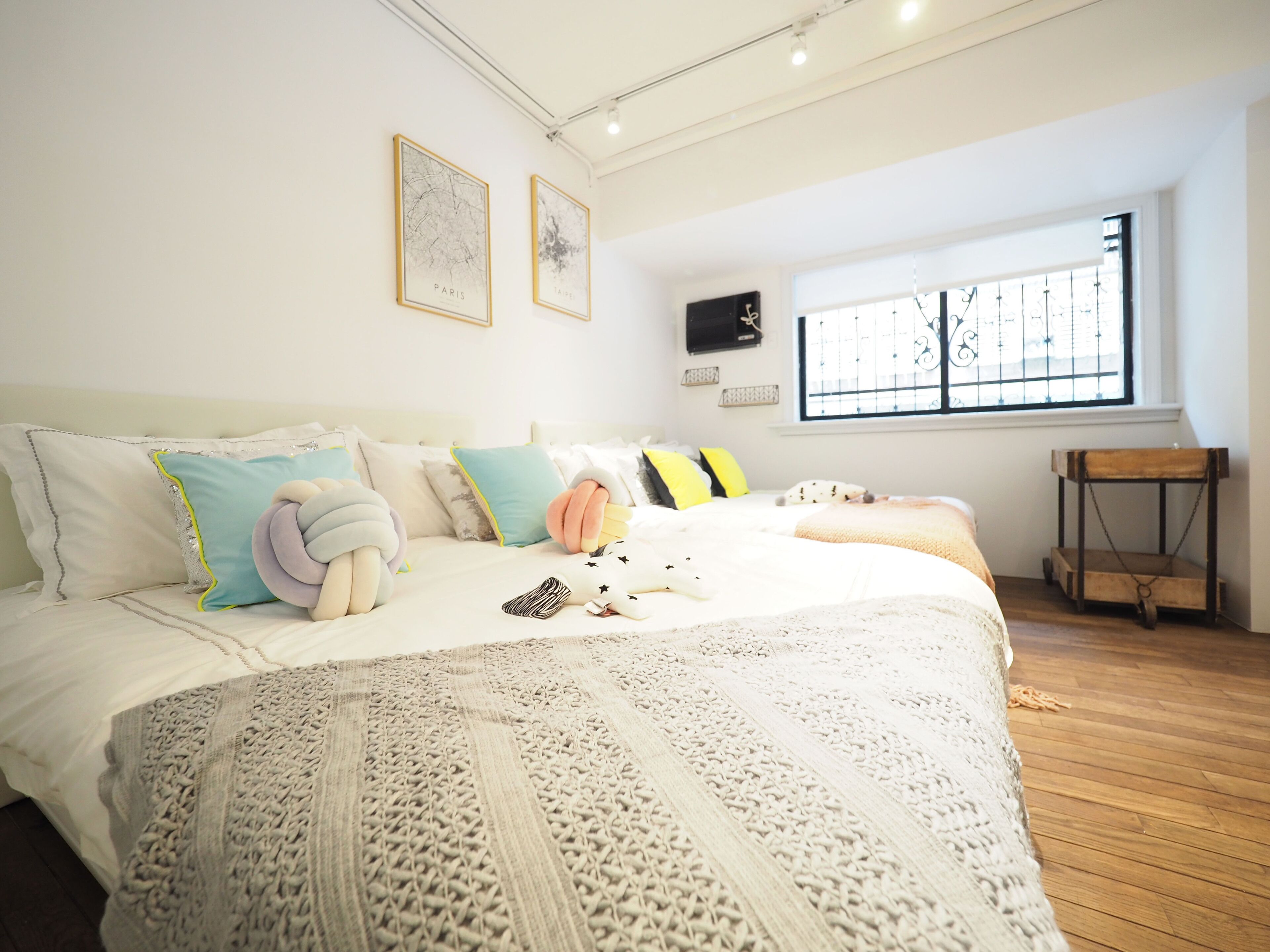 A bedroom with a large bed, multiple pillows, a knitted blanket, a window with bars, and a small wooden table.
