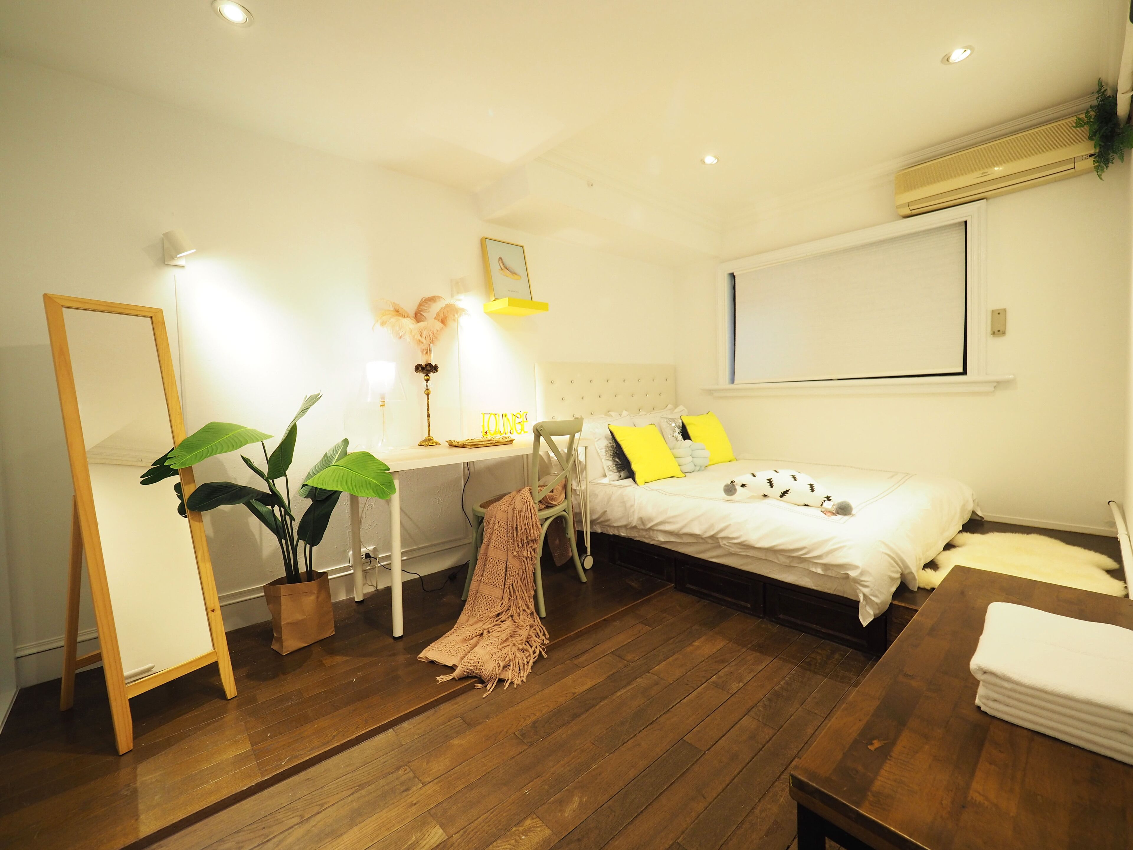 A bedroom with a bed, bedside table, mirror, desk, chair, and a potted plant.