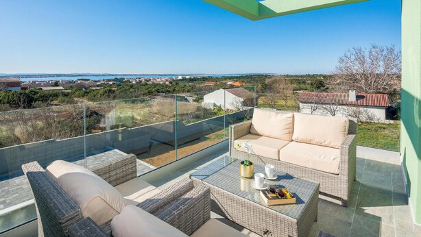 A balcony with wicker furniture, a glass table, and a view of a coastal town. - Beautiful home in Vrsi (Vrsi)