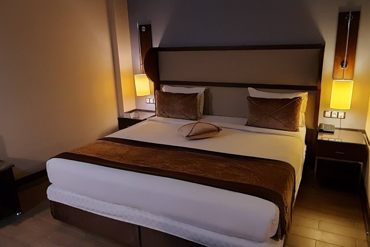 A hotel room with a large bed, two bedside tables, a nightstand, a wooden bench, and two wall-mounted lamps.