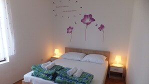 En pænt redt seng med et grønt og blåt ternet tæppe, hvide puder og to sammenrullede håndklæder. To sengeborde med lamper, samt en vægdekoration med lilla blomster og sommerfugle.