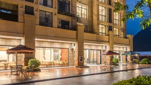 An outdoor area with tables and chairs under umbrellas, a building with multiple floors and balconies, and a wet pavement suggesting recent rain. - Meeteem Boutique Hotel (Baise)