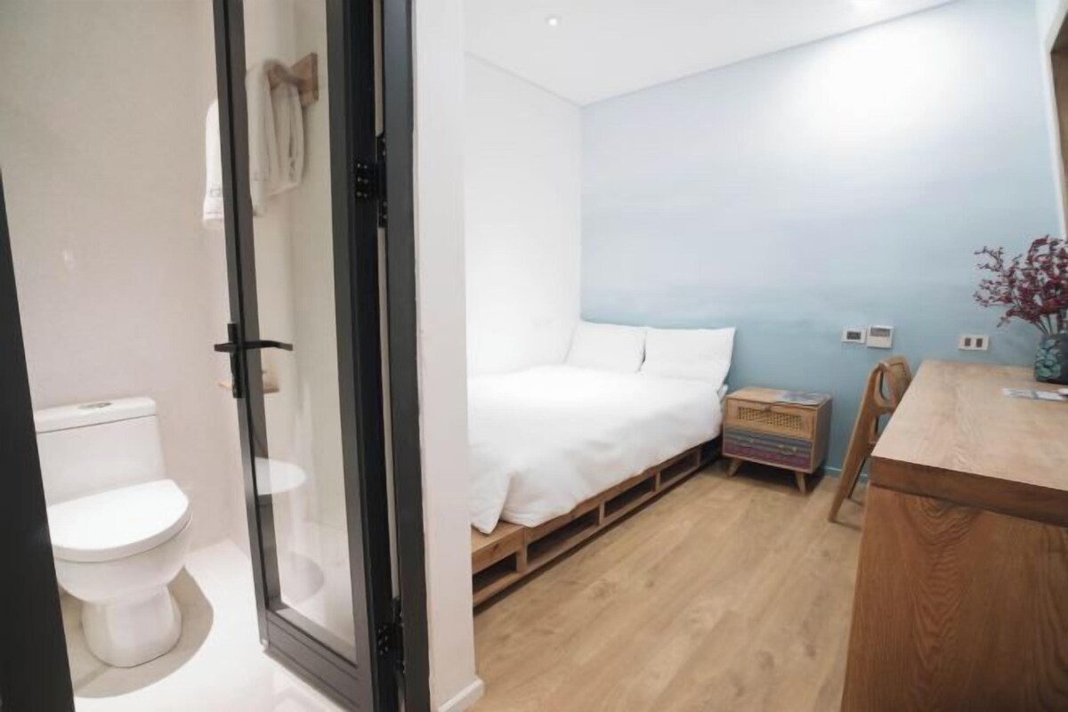 A bedroom with a bed, a wooden nightstand, a small table, and a wall-mounted shelf.