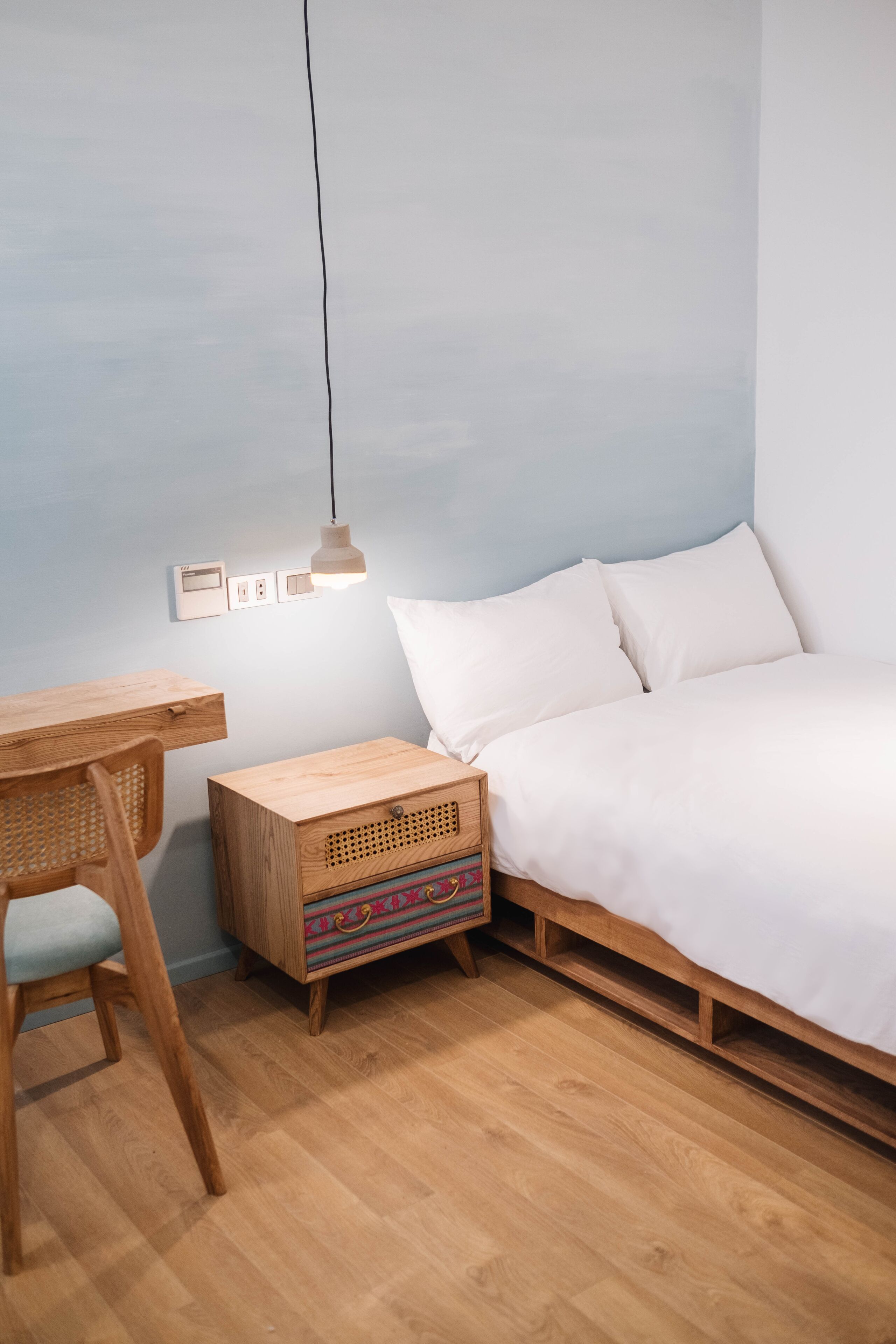 A bedroom with a wooden bed, a wooden nightstand with a colorful storage box, a wooden desk with a chair, and a hanging light fixture.