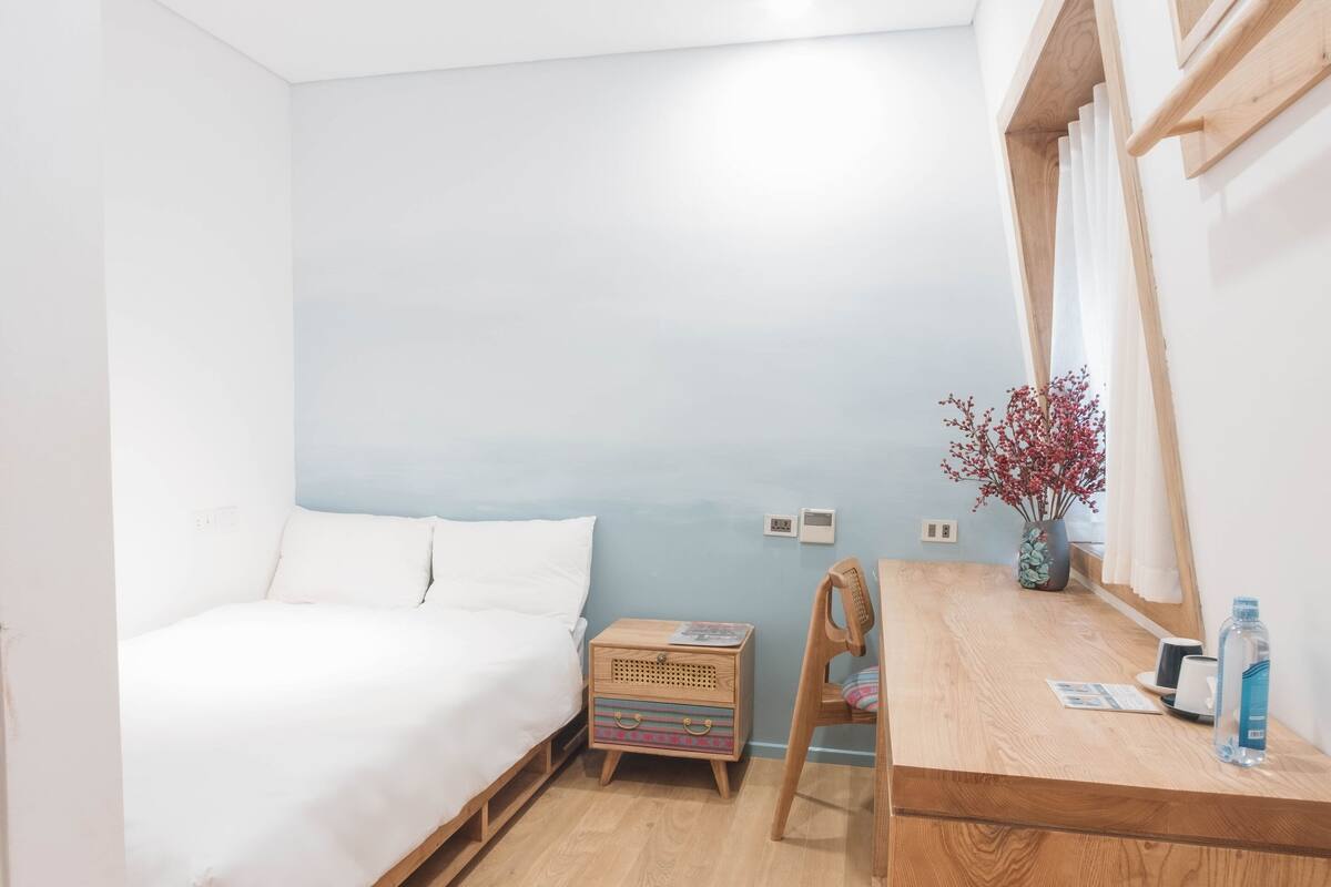 A bedroom with a bed, a wooden nightstand, a desk, a chair, a mirror, and a window with curtains.