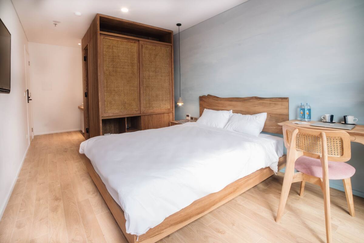 A bedroom with a wooden bed, a wooden wardrobe, a desk with a chair, and a wall-mounted shelf.