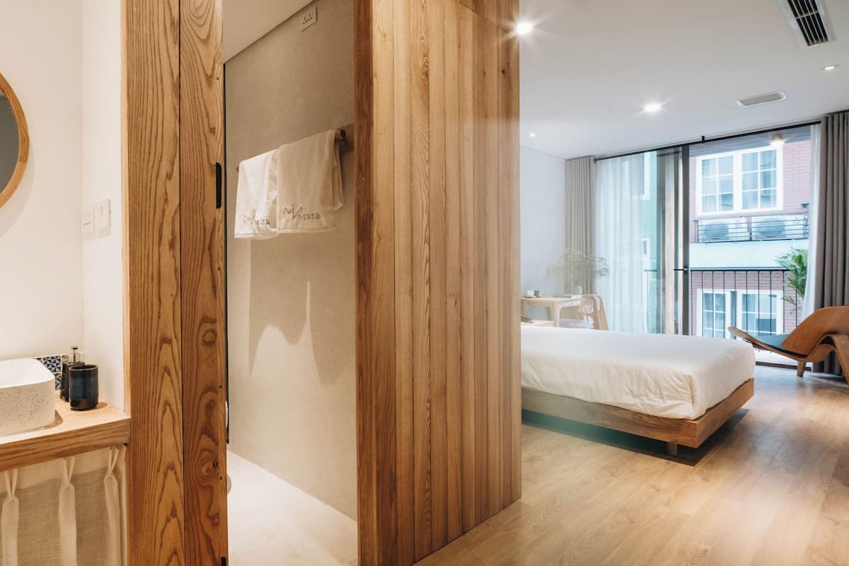 A modern hotel room with a wooden wardrobe, a round mirror, a large bed, and a view of the outside.