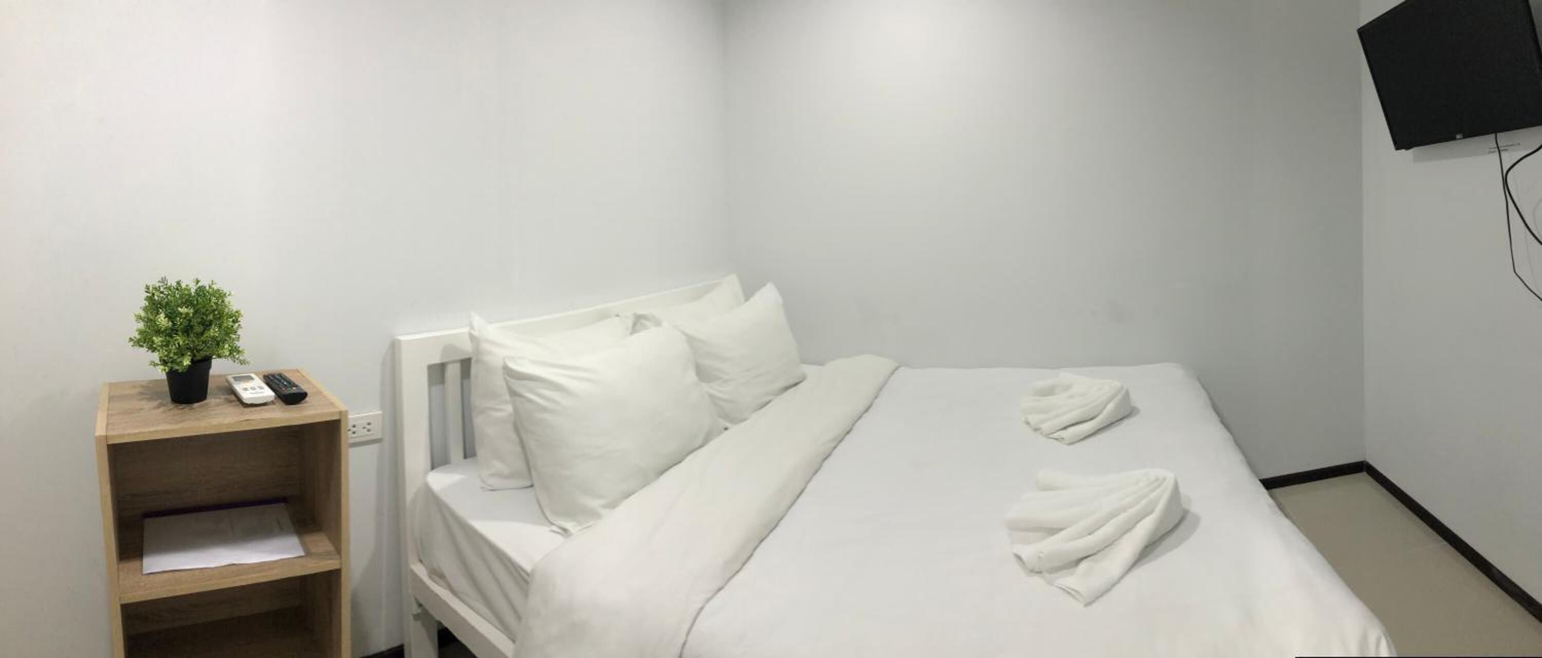 Une chambre d’hôtel avec un lit, du linge de lit blanc, une table de chevet avec une plante et un téléphone, et un téléviseur fixé au mur.