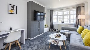 Een moderne woonkamer met een grijze bank, een wit bureau en een flatscreen televisie aan de muur.