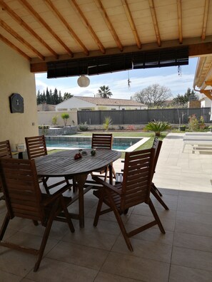 A covered patio with a round wooden table and chairs, a pool in the background, and a view of a residential area.