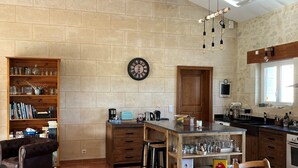A spacious kitchen with a large island, wooden beams, and a stone wall. There is a clock on the wall and a bookshelf filled with books and glassware.