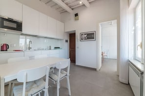 Une cuisine moderne avec des meubles blancs, une table à manger avec des chaises, une guitare encadrée accrochée au mur et une vue sur l’extérieur par une fenêtre.