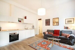 A modern living room with a grey sofa, a patterned rug, and a kitchen area with white cabinets and a built-in oven.