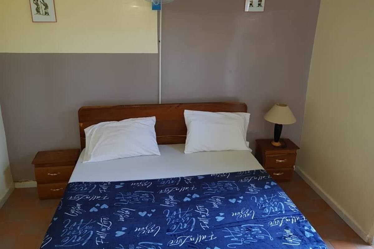 Un dormitorio con una cama, dos almohadas, una colcha azul con texto blanco, una mesita de noche con lámpara y un ventilador de pared.