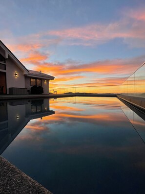 Een modern gebouw met een glazen balkon dat uitkijkt over een rustig zwembad dat de zonsondergang in de lucht weerspiegelt.