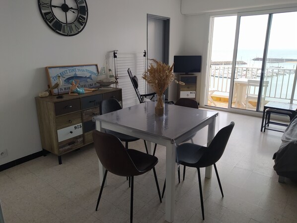A modern living room with a dining area, a large window with a view of the sea, a clock on the wall, and a flat-screen TV.
