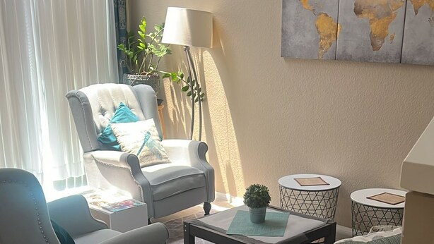 Una sala de estar con un sillón gris, un sofá oscuro, una mesa de centro y una mesita auxiliar con una planta. Hay un mapa mundial en la pared y una lámpara al lado del sillón.