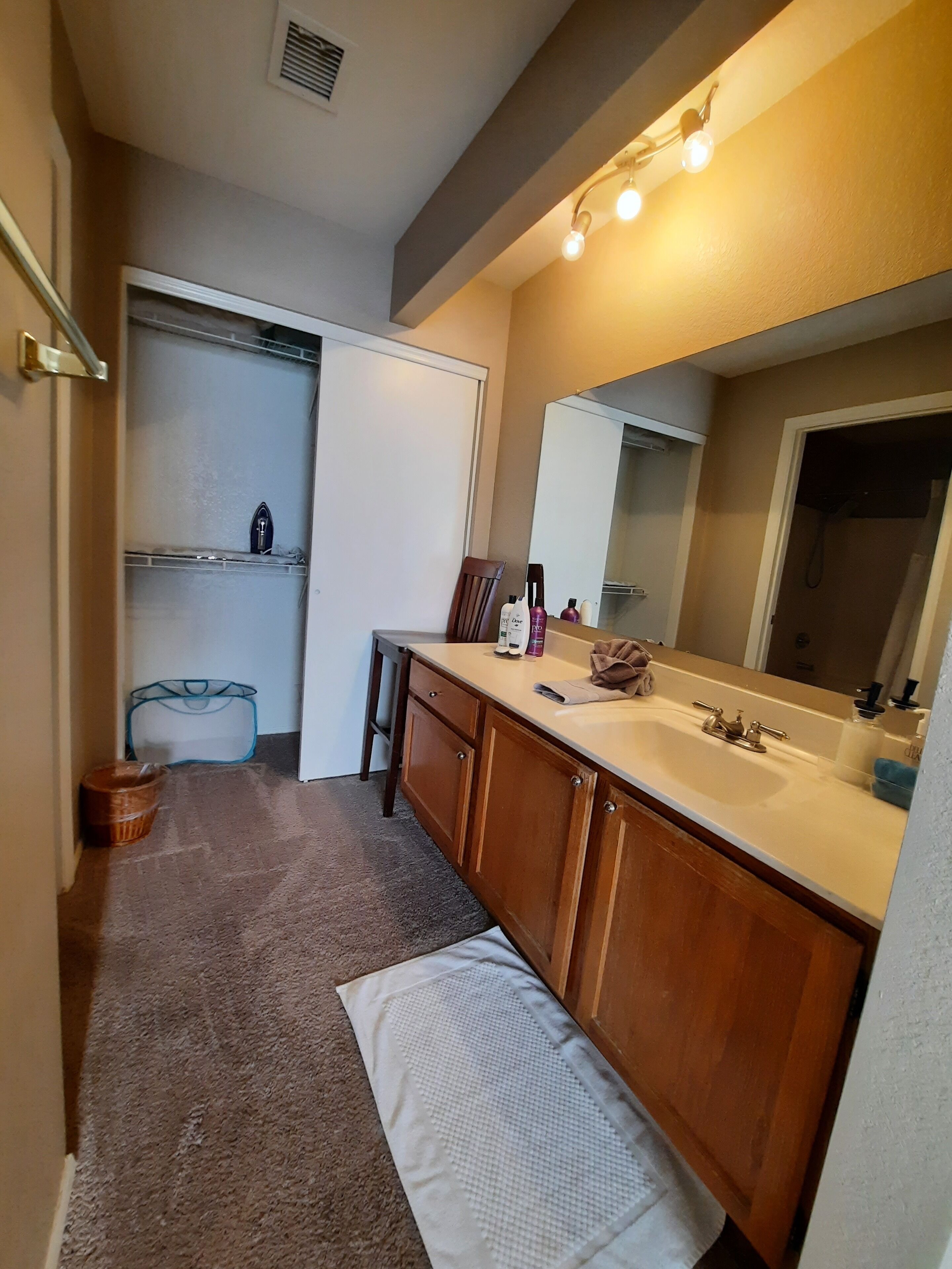 Une salle de bain avec un lavabo, un miroir et des placards de rangement. Il y a une serviette sur le sol et un panier près de la porte.