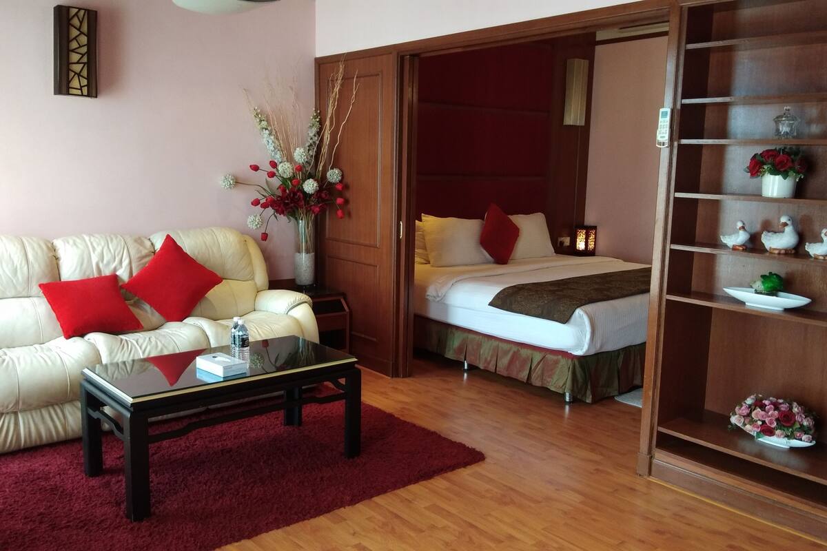 A hotel room with a sofa, a bed, a glass coffee table, and a wooden bookshelf.