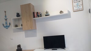 Een moderne woonkamer met een flatscreen televisie op een witte standaard, houten planken erboven met boeken en decoratieve objecten, en een klein beeldje op de vloer.