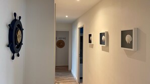 Un couloir avec un gouvernail de bateau sur le côté gauche, une tapisserie murale tissée et, sur le côté droit, des œuvres d’art représentant des coquillages encadrées.